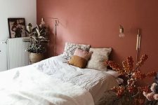 a bold bedroom with a terracotta accent wall, a bed with neutral bedding, gold sconces and potted plants