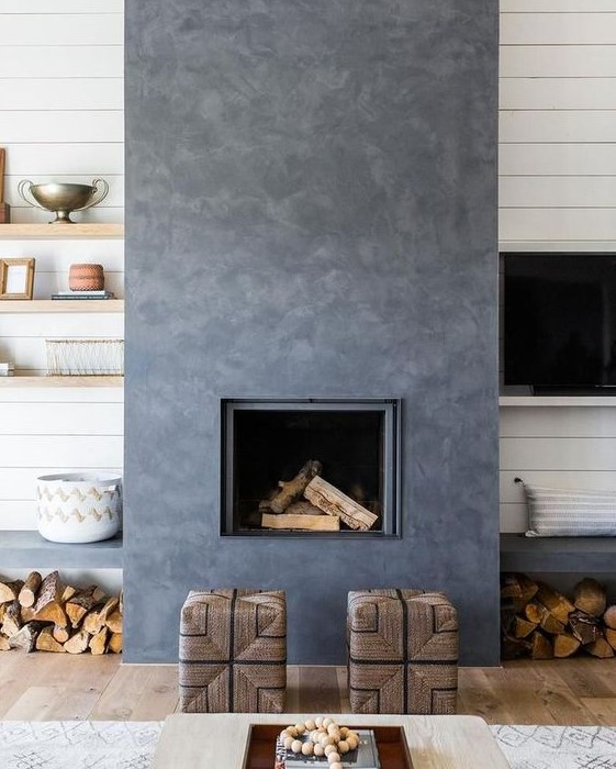 a catchy dark concrete fireplace with woven stools and benches with firewood under them is very cozy