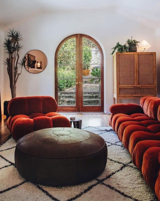 a catchy interior with bold rust colored seating furniture, a leather ottoman, a copper mirror and a rattan sideboard