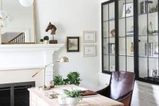 a chic home office with a built-in fireplace and a mantel, a mirror, greenery and art, a light stained desk, a leather chair and touches of brass