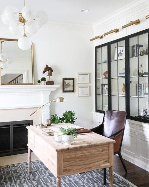a chic home office with a built in fireplace and a mantel, a mirror, greenery and art, a light stained desk, a leather chair and touches of brass