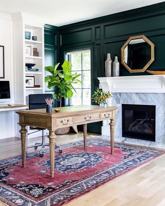 a chic home office with black paneling, niche shelves, a fireplace clad with marble, a rustic vintage desk, a boho rug and potted plants