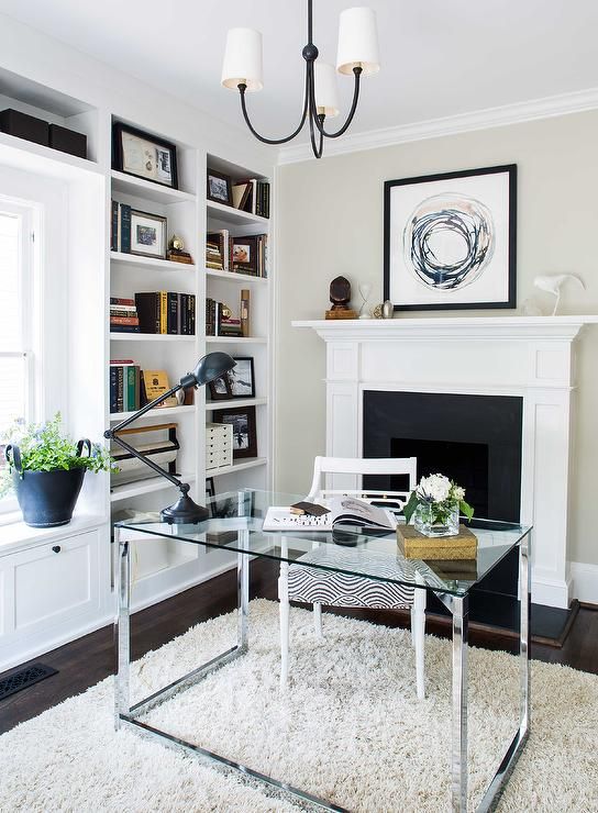 a chic modern neutral home office with built in bookshelves, a fireplace, a glass desk, a neutral rug and a stylish chandelier