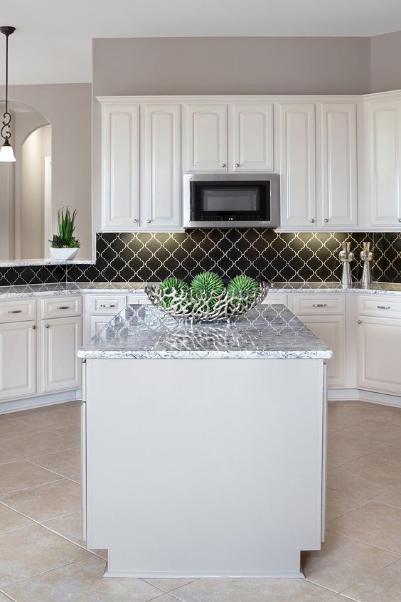 a chic modern white kitchen with shaker cabinets, white stone countertops, a black arabesque tile backsplash and greenery