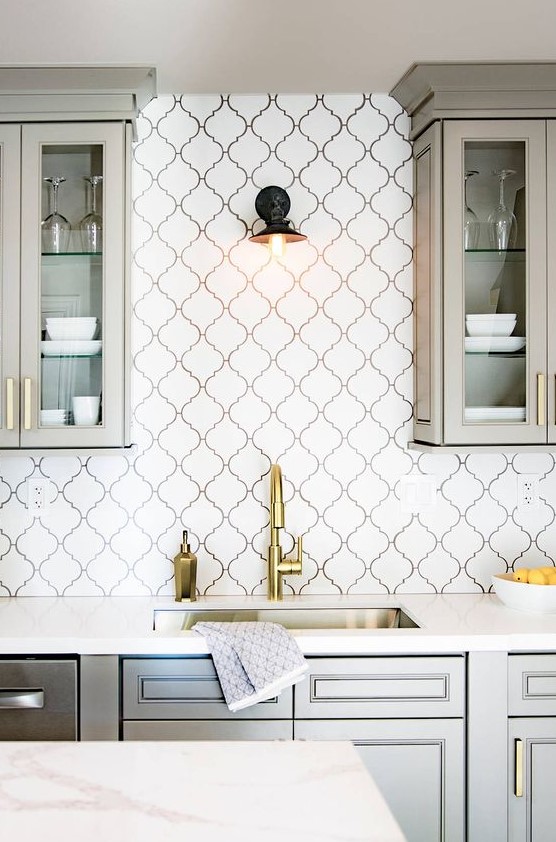 a contemporary light grey kitchen with glass cabinets, a white arabesque tile backsplash accented with black grout and gold fixtures