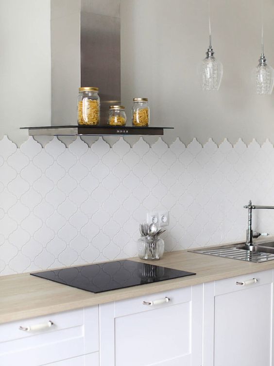 a contemporary white kitchen with neutral butcherblock countertops, sleek modern appliances, an arabesque tile backsplash and pendant lamps