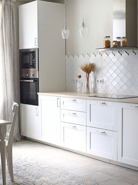 a contemporary white kitchen with shaker cabinets, butcherblock countertops, a white arabesque tile backsplash, pendant bulbs is chic