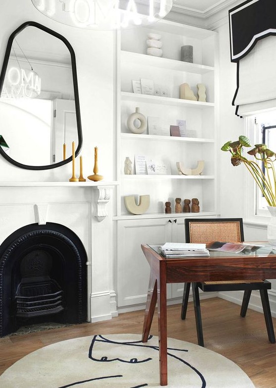 a creative and refined home office with a built-in storage unit, a metal fireplace, an asymmetrical mirror in a black mirror, black and white curtains, a rich stained desk and a rattan chair