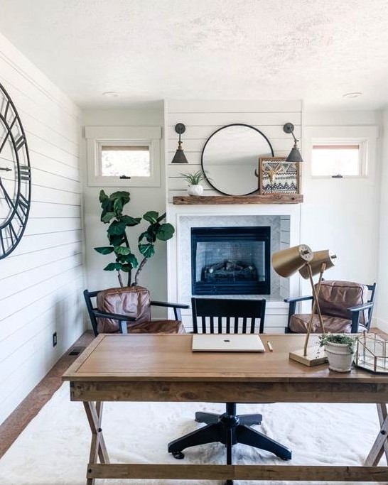 a farmhouse home office with white planked walls, a built in fireplace, a trestle desk, leather chairs and a black one, some decor and plants