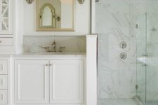 a glam vintage-inspired bathroom with white marble and arabesque tiles, white furniture, a shower space, a mirror in a metallic frame