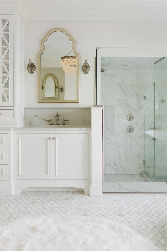a glam vintage inspired bathroom with white marble and arabesque tiles, white furniture, a shower space, a mirror in a metallic frame