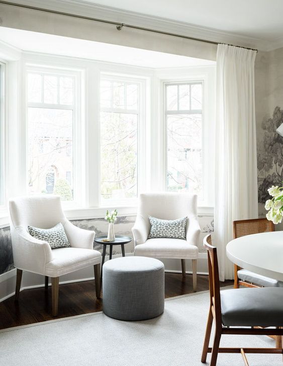a large bay window turned into a sitting zone with white chairs, printed pillows, a side table and a grey pouf