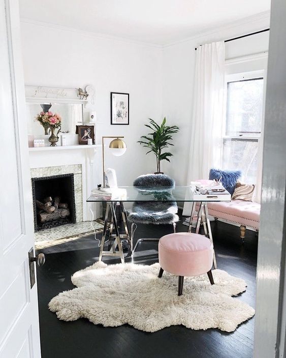 a light filled home office with a fireplace, a glass trestle desk, a pink sofa and a pouf, potted plants, brass lamps looks cute and cool