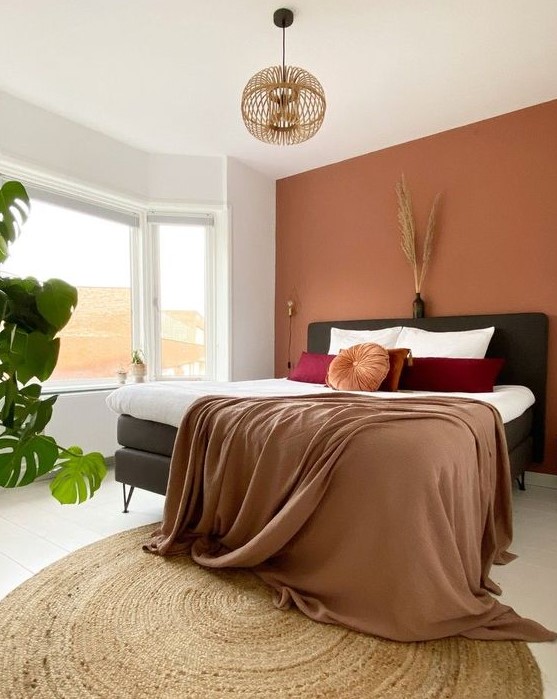 a lovely bedroom with a boho feel, a terracotta accent wall, a black bed, bold bedding, a wooden pendant lamp and some plants
