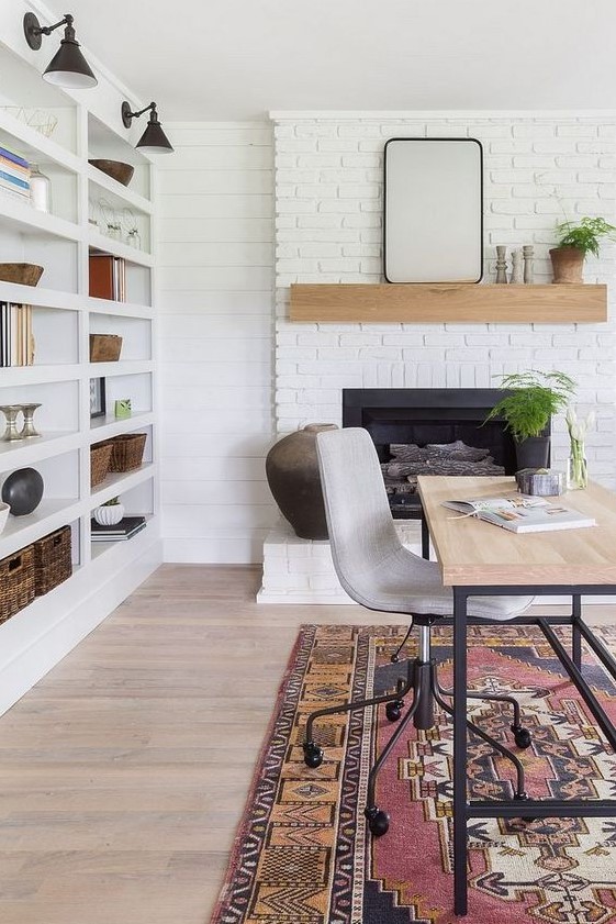 a lovely modern farmhouse home office with a white brick fireplace, a mantel, a large shelving unit with sconces, a delicate desk and printed rug