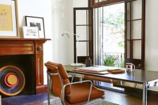 a mid-century modern home office with an entrance to the blacony, a non-working fireplace, a desk, an orange leather chair, a printed rug and chic artworks