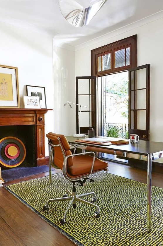 a mid-century modern home office with an entrance to the blacony, a non-working fireplace, a desk, an orange leather chair, a printed rug and chic artworks