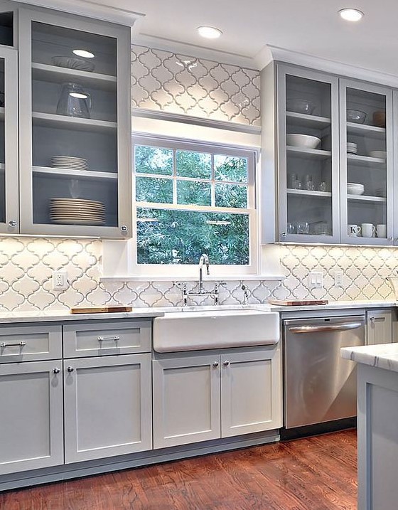 a modern light grey kitchen with shaker cabinets and glass ones, with a white wall clad with white arabesque tiles and white stone countertops