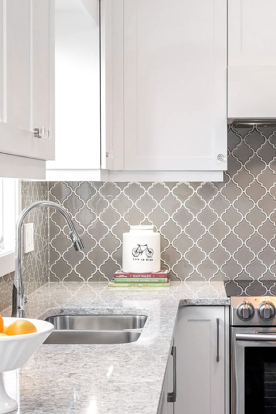 a modern white kitchen with a white stone countertop and a grey arabesque tile backsplash, with stainless steel fixtures