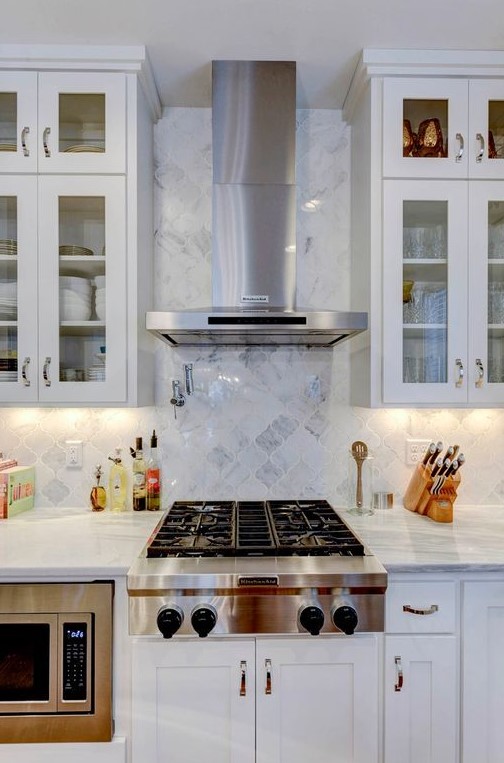 a modern white kitchen with upper glass cabinets, with a white marble arabesque tile backsplash and built-in appliances is amazing