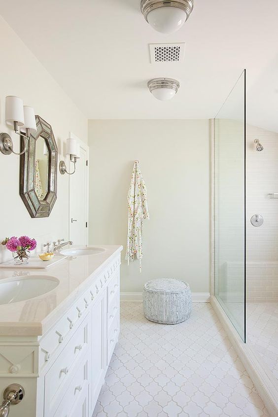 a neutral bathroom with tan walls, a white arabesque tile floor, a white vanity and a chic geometric mirror plus lamps