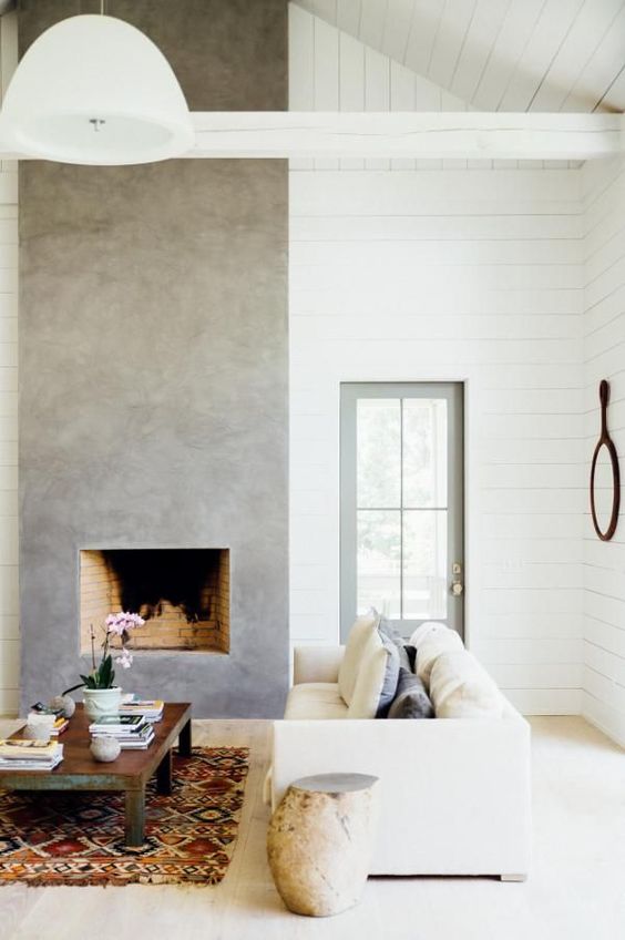 a neutral farmhouse living room with a concrete fireplace, neutral seating furniture, a low coffee table, a printed rug and a pendant lamp