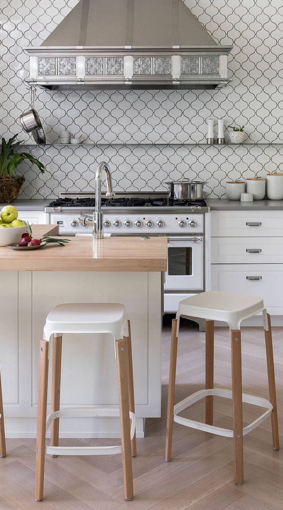 a pretty and catchy white kitchen with a white arabesque tile backsplash, grey stone countertops, stainless steel appliances and an ornated hood