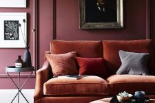 a refined and moody living room with a burgundy paneled wall, a gallery wall, a rust-colored sofa and an ottoman plus colorful pillows