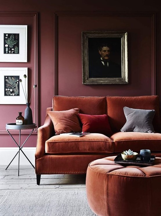 a refined and moody living room with a burgundy paneled wall, a gallery wall, a rust colored sofa and an ottoman plus colorful pillows