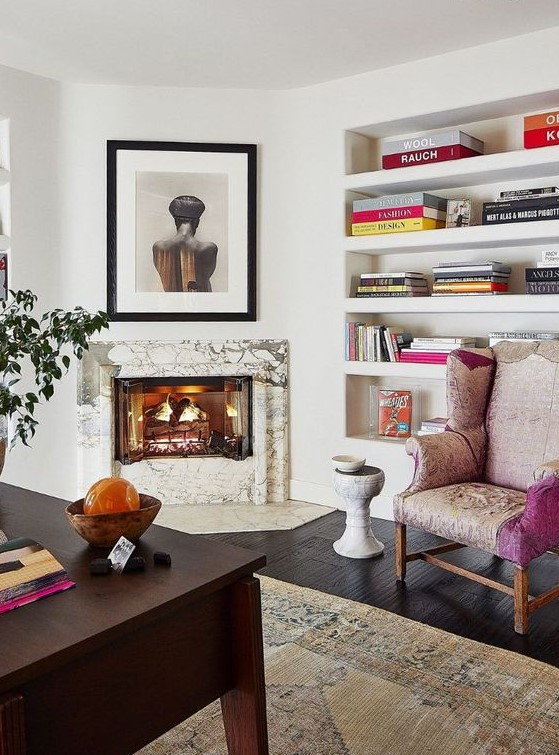 a refined home office with a marble clad fireplace, built-in shelves, a dark stained desk, a pink chair and some art and bright books