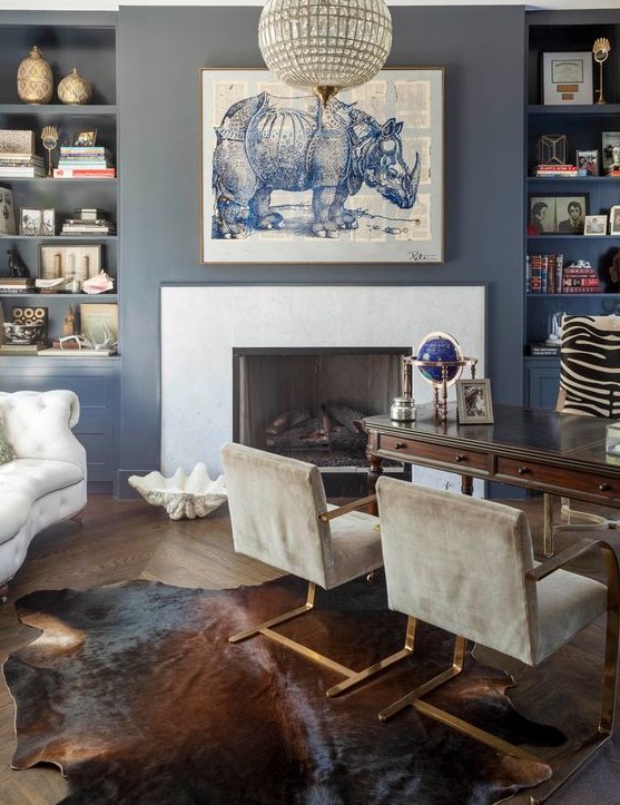 a refined home office with blue walls, built-in shelves, a fireplace, a vintage dark stained desk, grey chairs and a creamy sofa plus art