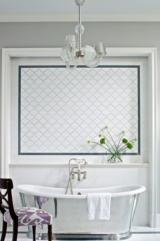 a refined vintage bathroom with grey walls, a white arabesque tile accent, a shiny polished bathtub and a chic crystal chandelier