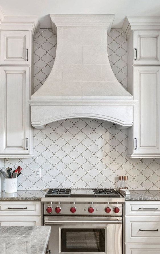 a refined vintage white kitchen with shaker style cabinets, a white arabesque tile backsplash and a vintage hood plus built-in appliances