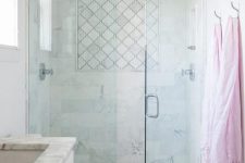 a small and chic neutral bathroom with white marble and arabesque tiles, a small shower enclosed in glass, a white vanity and stainless steel fixtures