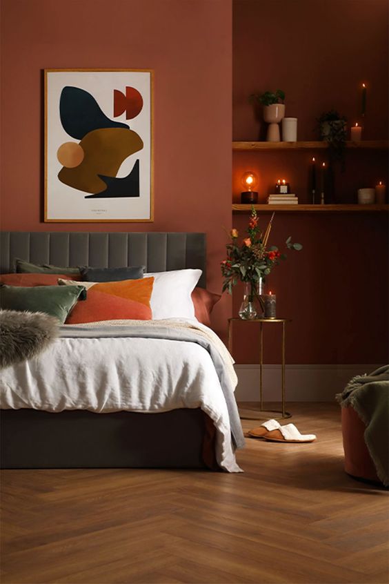 a soothing bedroom with terracotta walls, a grey upholstered bed with orange and white bedding, wall-mounted shelves and some lovely decor
