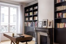a sophisticated home office with black built-in bookcases, a chic fireplace with a wooden surround, a mid-century modern desk and a black chair