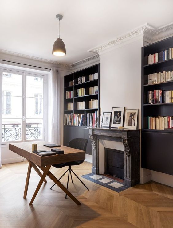 a sophisticated home office with black built-in bookcases, a chic fireplace with a wooden surround, a mid-century modern desk and a black chair