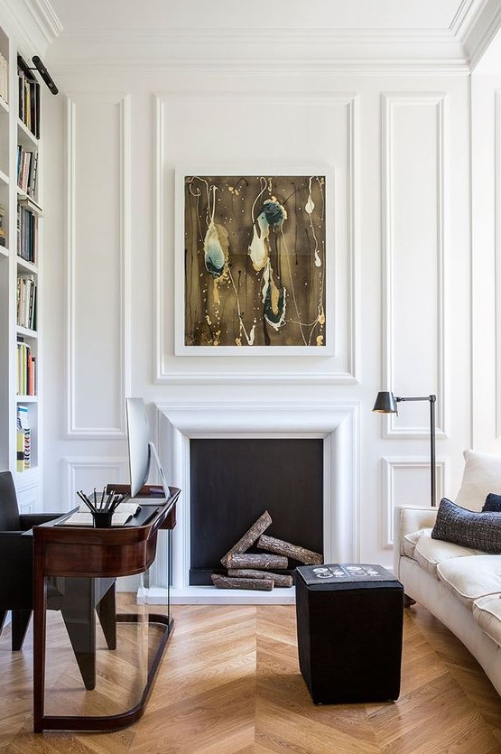 a sophisticated home office with white paneling, a non-working fireplace, a dark-stained rounded desk, a black pouf and a creamy sofa is wow