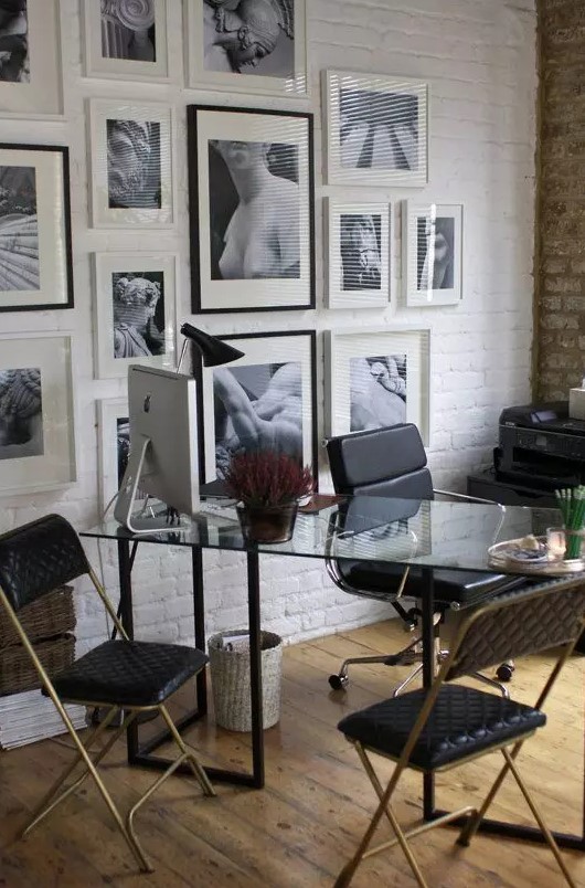 a stylish home office with a white brick accent wall with a gallery wall, a glass desk, black leather chairs and some potted plants