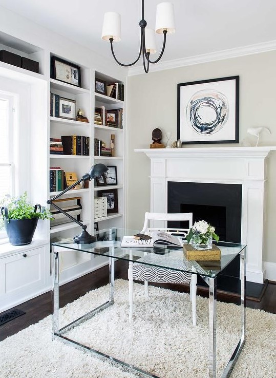 a stylish home office with built-in shelves, a glass desk, a fireplace behind it, a windowsill seat and potted plants and blooms
