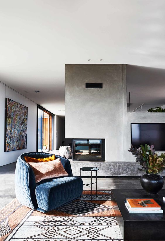 a stylish mid century modern living room with a concrete fireplace, a blue quilted chair, a printed rug and a bold artwork