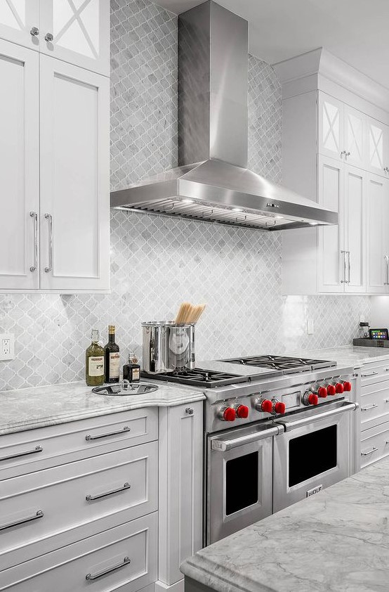 a stylish white kitchen with shaker style cabinets, grey stone countertops, a grey marble arabesque tile backsplash plus cool appliances