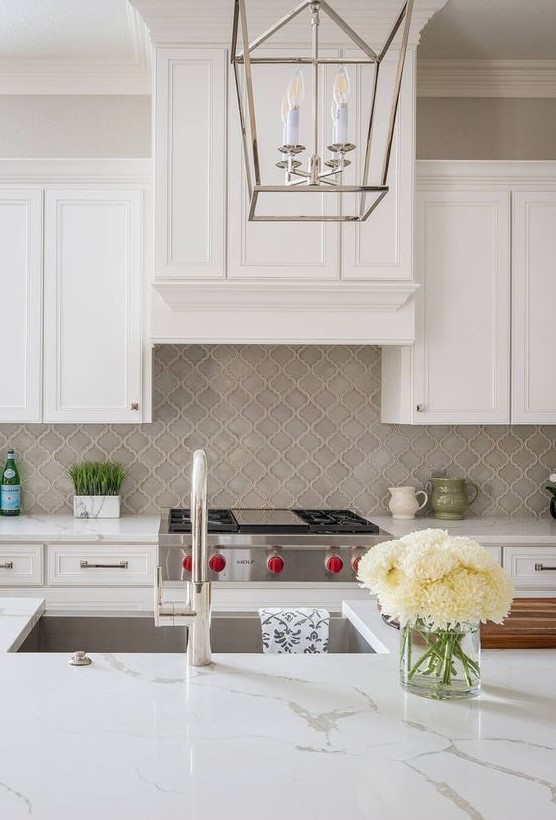 a vintage and refined white kitchen with shaker style cabinets, a grey arabesque tile backsplash, white stone countertops and chromatic appliances