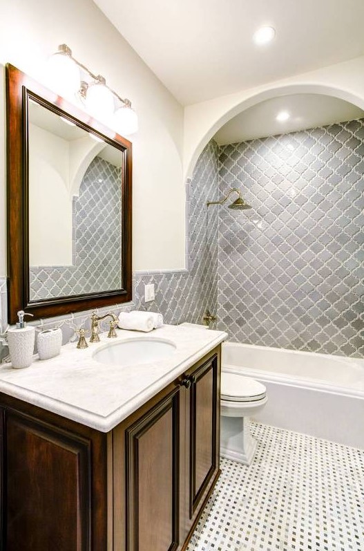 a vintage bathroom with grey arabesque tiles and mosaic ones on the floor, a stained vanity and a mirror in a stained frame