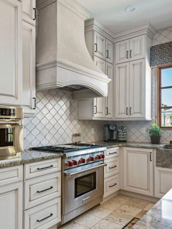 a vintage neutral kitchen with shaker cabinets, a grey stone countertop and a neutral arabesque tile backsplash, a large hood