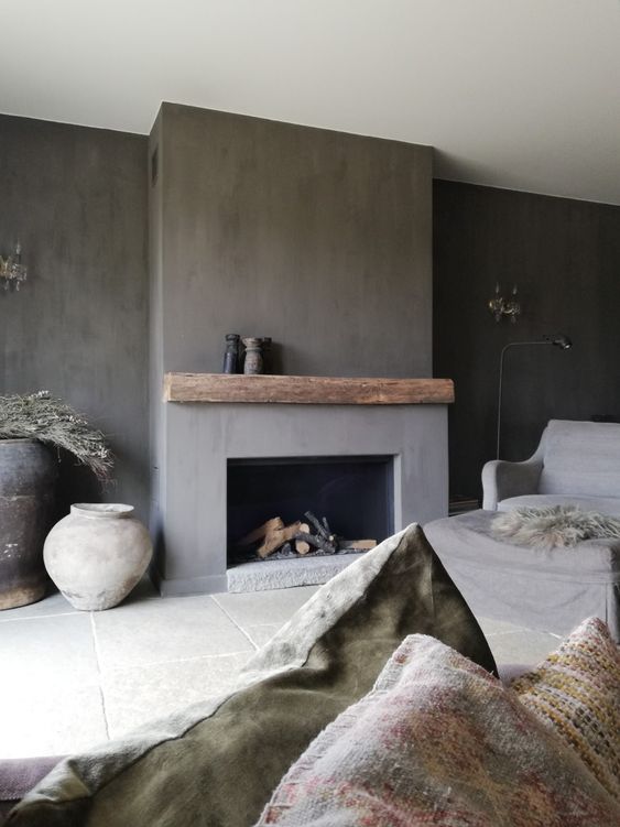 a wabi-sabi living room with concrete walls and a fireplace, a sofa with printed textiles, large vases and firewood in the fireplace