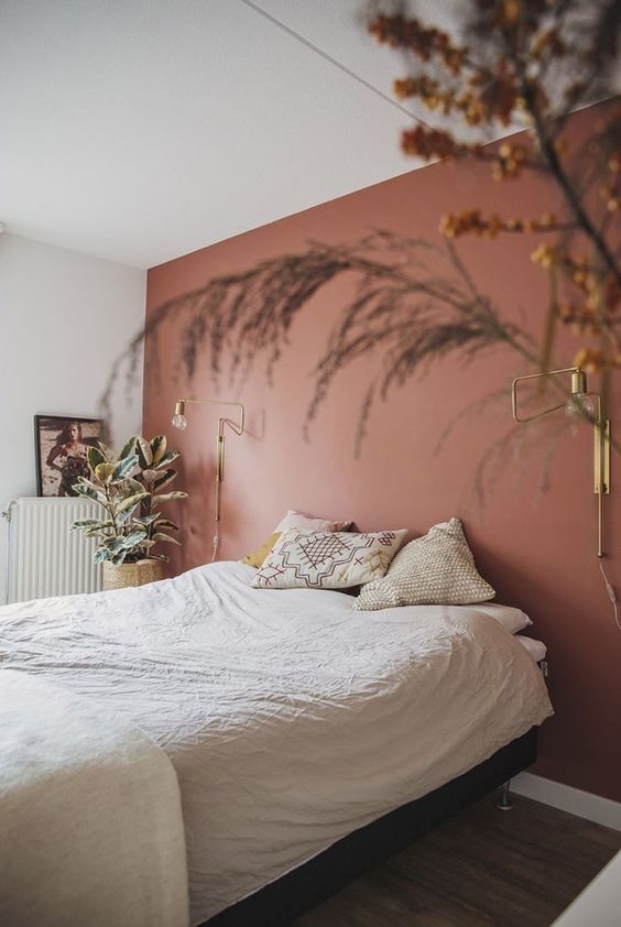 a welcoming bedroom with a terracotta accent wall, a bed with neutral bedding, gold sconces and potted plants and grasses
