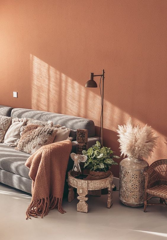 a welcoming boho living room with a terracotta accent wall, a grey sofa with pillows, a carved boho side table, pampas grass and a floor lamp