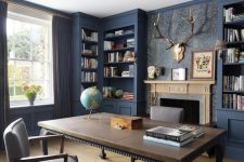 a whimsical home office with built-in blue bookcases, a fireplace, a vintage desk, a taupe chair and dark curtains plus antlers