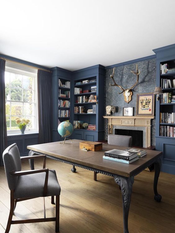 a whimsical home office with built-in blue bookcases, a fireplace, a vintage desk, a taupe chair and dark curtains plus antlers